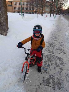 Un enfant de 7 ans pose avec son vélo sur un trottoir enneigé au centre de la photo. Il est habillé d'un manteau d'hiver et de pantalons de neige orange, de mitaines noires, de bottes noires et d'un foulard. Il porte un casque noir et des lunettes de ski bleues, ainsi qu'un sac à dos. L'enfant a ses deux mains sur son guidon. À sa gauche, il y a un parc urbain enneigé devant un bâtiment. À sa droite, il y a le trottoir sur lequel il reste une mince couche de neige, puis une grande rue.