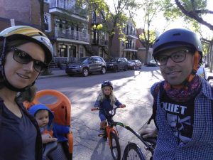 La photo est un égoportrait qui montre une famille à vélo. Il y a quatre personnes qui posent devant une rue résidentielle à Montréal. Il fait soleil et c'est l'été. À gauche, une femme porte des lunettes de soleil et un casque de vélo. Derrière elle, un petit enfant en veste bleue est assis dans un siège orange fixé sur le vélo. À droite du petit enfant, il y a un enfant aux longs cheveux blonds et portant un casque assis sur un petit vélo. Le petit vélo est attaché par une barre à un vélo d'adulte. À la toute droite de la photo, un homme portant un casque et des lunettes sourit. Il porte une chemise à carreaux ouverte sur un t-shirt noir sur lequel on lit le mot «quiet» en blanc.